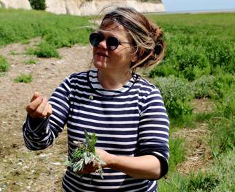 A woman wearing sunglasses and a striped t-shirt holding a bunch of herbs foraged from the shore with the white cliffs in the background.