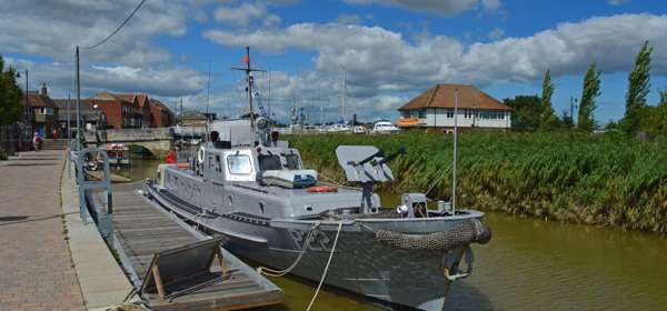 A grey patrol boat on the river next to a boarding quay with buildings in the distance