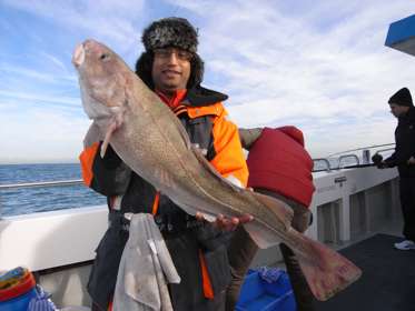 A man wearing a black and orange jacket and a fur hat holding a large fish aboard a fishing boat at sea.