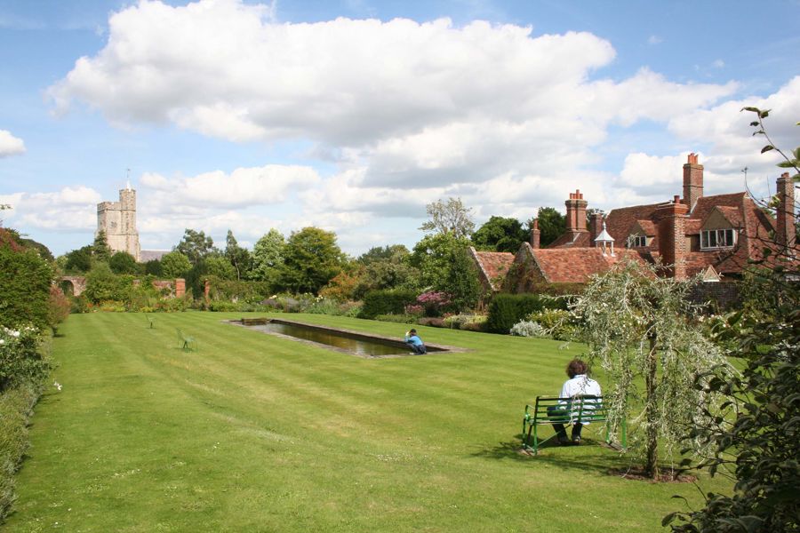 A green lawn with a long rectangular pond, a person sitting on a bench, a red-rooved house to the right and a church tower in the distance.