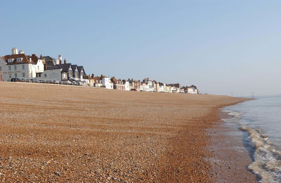 A shingle beach with waves lapping at the shore and a row of houses and hotels on the seafront in the distance. 