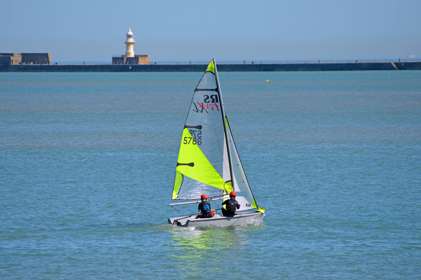 Two people wearing red protective helmets on a sailing dingy with a fluorescent yellow sail in Dover Harbour.