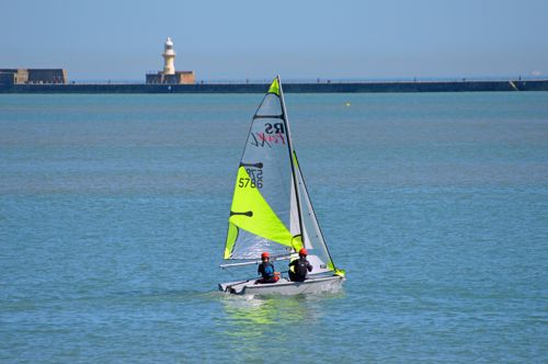 Two people wearing red protective helmets on a sailing dingy with a fluorescent yellow sail in Dover Harbour.