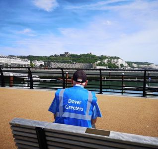 A man wearing a blue high-vis tabard with the words Dover Greeters on the back sitting on a bench in the foreground, with Dover seafront and castle in the distance