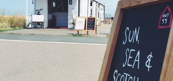A chalkboard sign saying 'Sun, Sea & Scotch Eggs' pointing at a beach hut cafe, Hut 55.