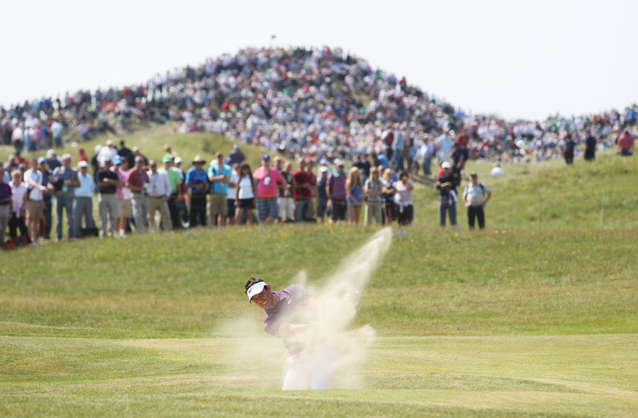 A golfer in a bunker with sand spraying after a shot; a crowd of spectators in the background. 
