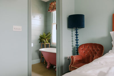 A pale blue bedroom with pink armchair, dark blue standard lamp and doorway to bathroom with pink roll-top bath.