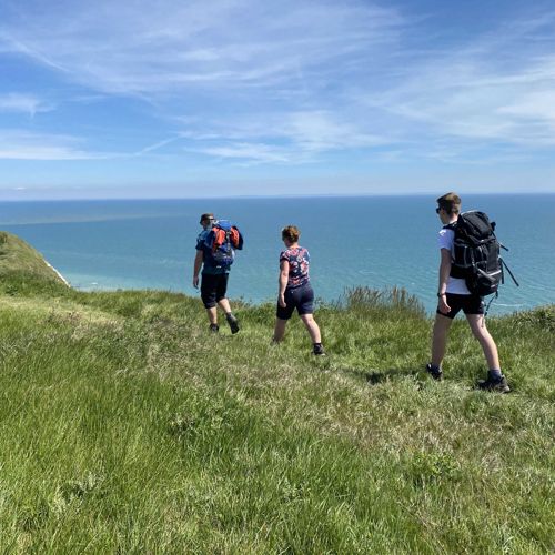 Three walkers carrying rucksacks walking along the cliff edge with blue sea and blue sky in the background.