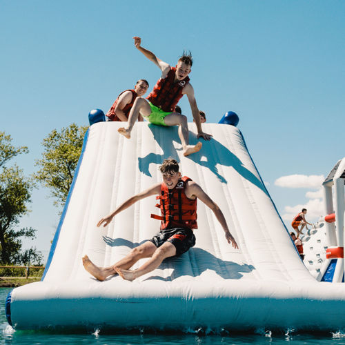 Young people wearing red life jackets and swimsuits sliding down a giant, floating, inflatable slide into blue water.