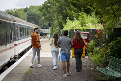 Four people on a station platform with a train to the left of the image and a green fence and trees to the right.