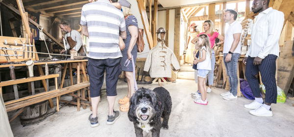 A group of people watching a blacksmith at work at Sandwich Medieval Centre with a grey and white dog in the foreground looking at the camera.