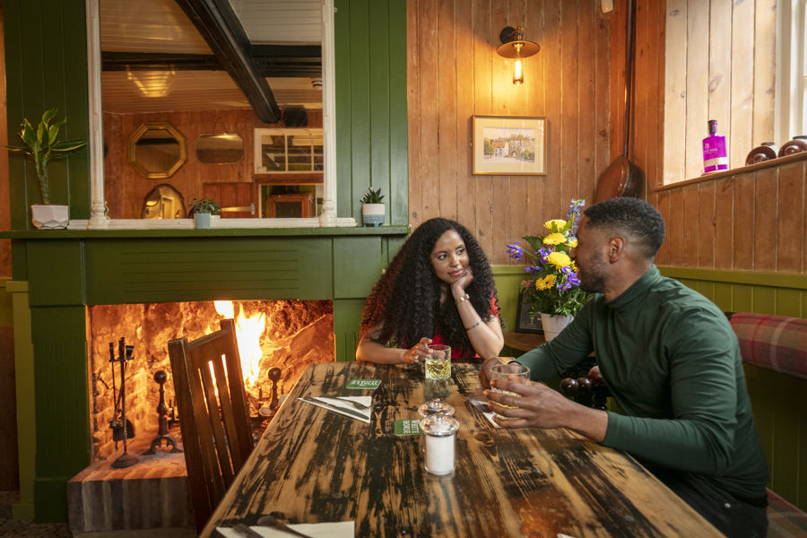 a couple sitting at a wooden table in a pub with a roaring fire to the left of the picture