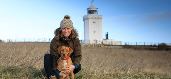 A woman wearing a coat and bobble hat crouched with a brown dog with a red collar both looking at the camera. The South Foreland Lighthouse and a blue sky behind.