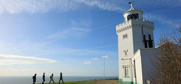 4 people approaching a white painted lighthouse on foot, across a green lawn with the sea just visible in the background.