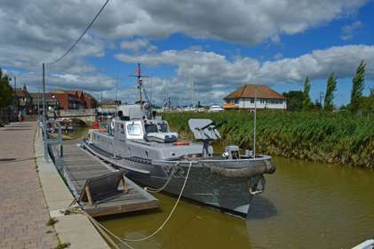 A grey patrol boat on the river next to a boarding quay with buildings in the distance