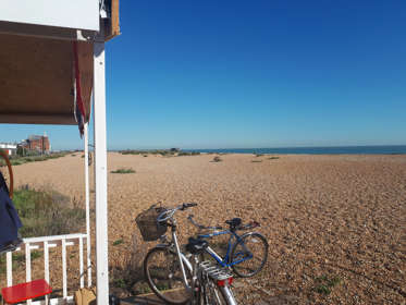 Two bicycles parked up next to a beach hut on a shingle beach with the sea and blue sky in the distance. 