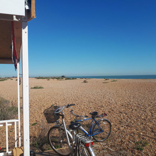 Two bicycles parked up next to a beach hut on a shingle beach with the sea and blue sky in the distance. 