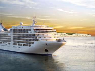 Large white cruise ship on a calm sea with the White Cliffs in the background and a yellow tint in the sky.
