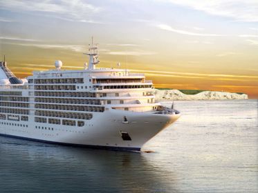 Large white cruise ship on a calm sea with the White Cliffs in the background and a yellow tint in the sky.