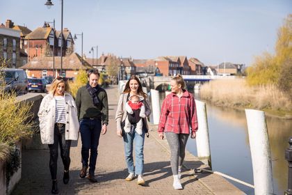 Three women and one man walking towards the camera along the quay at Sandwich next to the river with buildings in the background