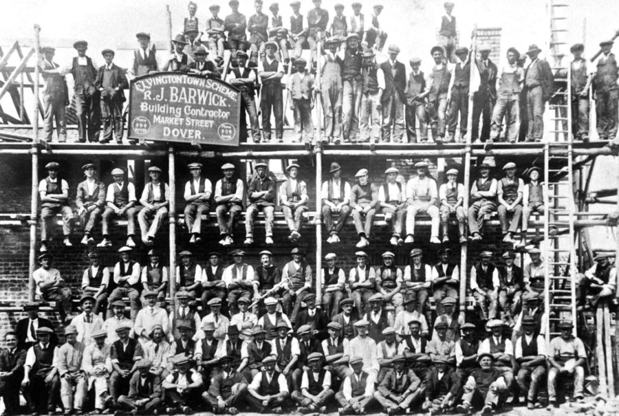 B&W photo of about one hundred men sat on a building site.