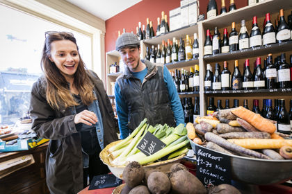 A man and woman inside a food shop with bottles of wine lining the wall behind and piles of vegetables in the foreground.