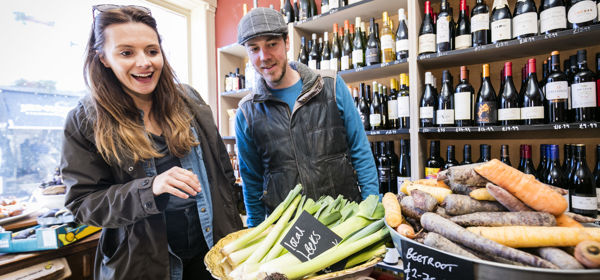 A man and woman inside a food shop with bottles of wine lining the wall behind and piles of vegetables in the foreground.