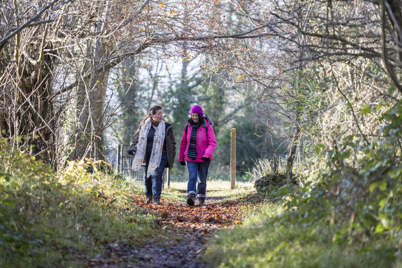 Two women walking towards the camera along a woodland path, one wearing a pink coat and hat, both wearing walking boots. The trees are bare and there are leaves on the path.