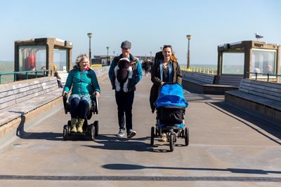Three people coming on Deal Pier, one in a wheelchair, one pushing a buggy and one carrying a baby in a papoose.