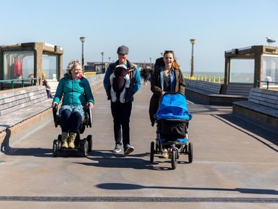 Three people coming on Deal Pier, one in a wheelchair, one pushing a buggy and one carrying a baby in a papoose.