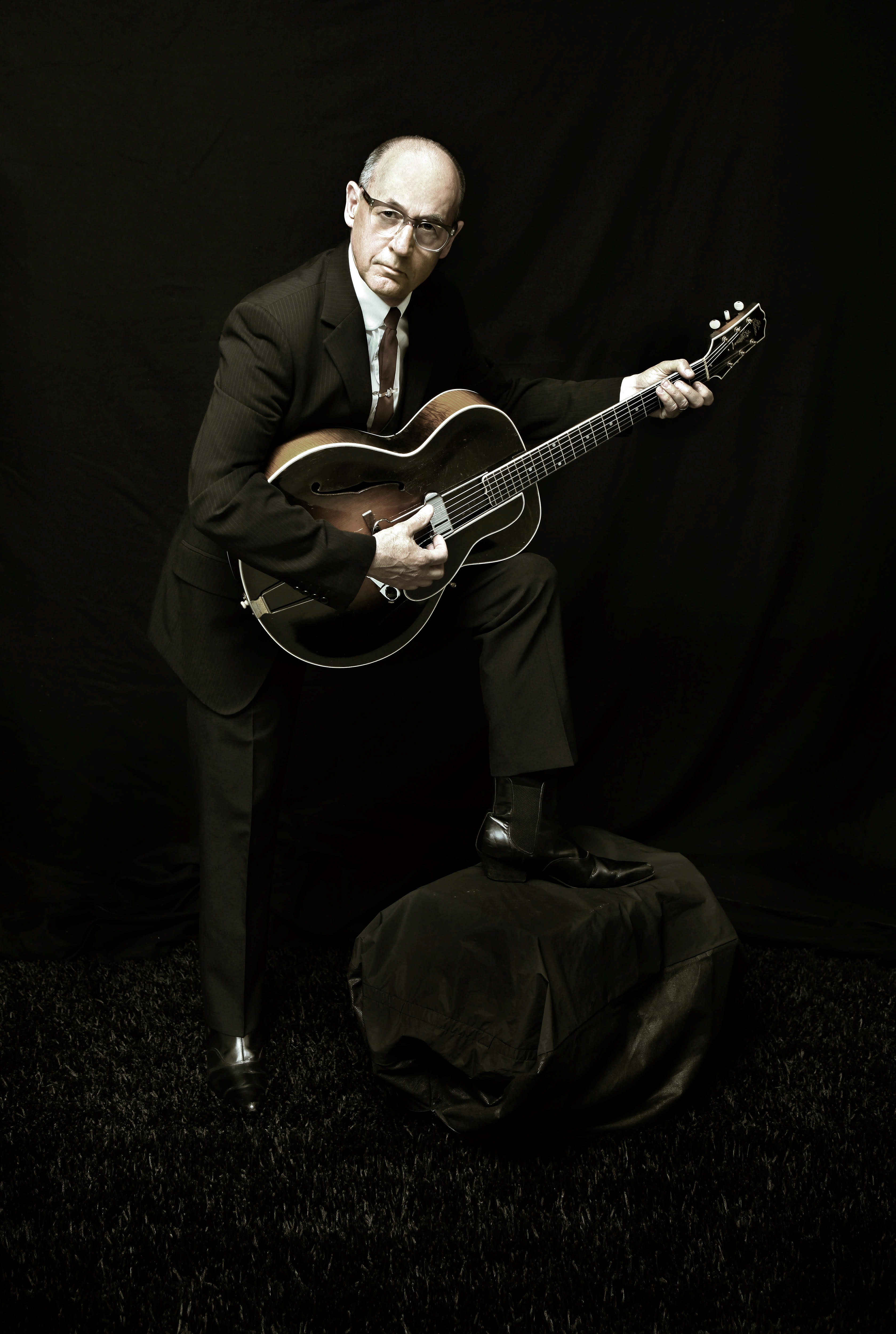 A sepia image of Andy with a foot raised on a stool strumming a guitar