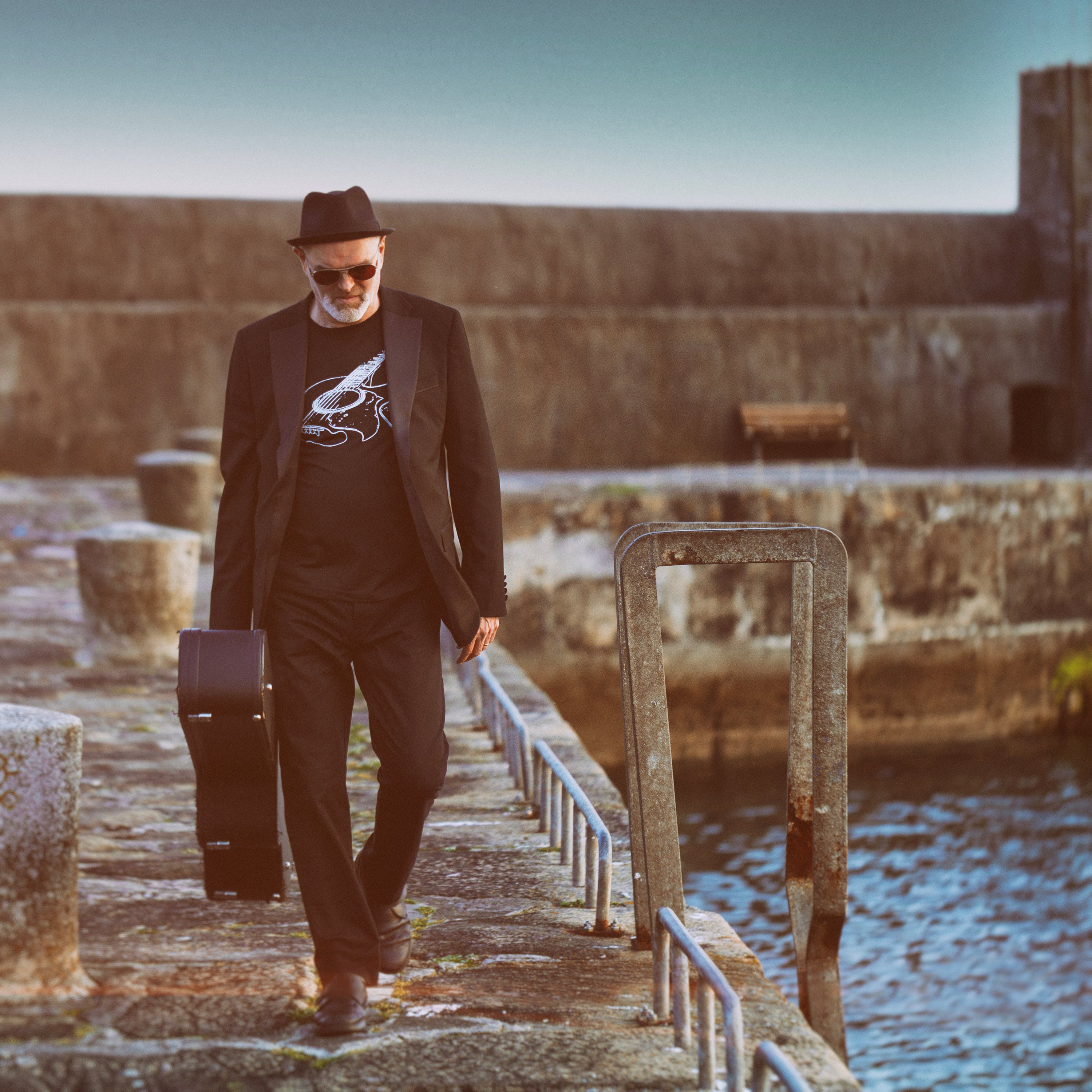 Ben walking along the docks dressed from head to toe in black carrying his guitar case