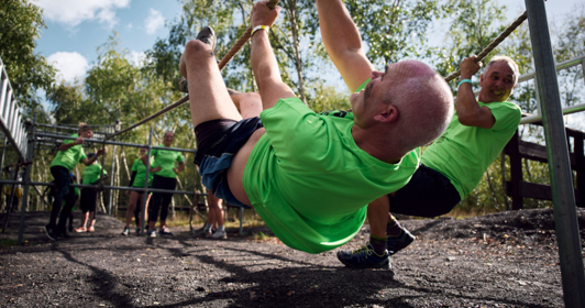 A group wearing green t-shirts navigate an outdoor obstacle course. The man in the foreground is hanging upside down from a rope.