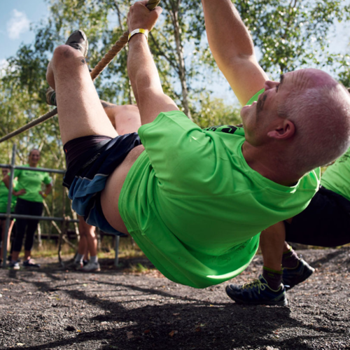 A group wearing green t-shirts navigate an outdoor obstacle course. The man in the foreground is hanging upside down from a rope.