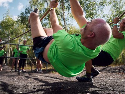 A group wearing green t-shirts navigate an outdoor obstacle course. The man in the foreground is hanging upside down from a rope.
