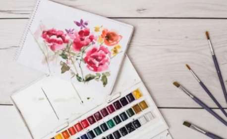 Sketch pad with painting of colourful flowers next to a palette of watercolour paints and brushes
