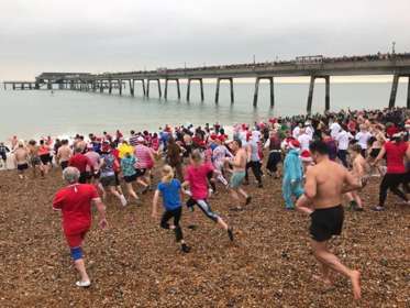 A crowd of people in colourful costumes and swimwear running down a shingle beach towards the sea with Deal Pier the background full of spectators.