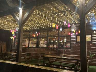 Wooden picnic tables under a wooden pergola covered in fairy lights and colourful lanterns lit up at night. 