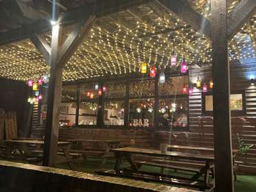 Wooden picnic tables under a wooden pergola covered in fairy lights and colourful lanterns lit up at night. 