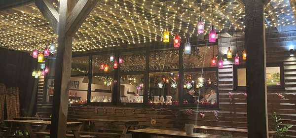 Wooden picnic tables under a wooden pergola covered in fairy lights and colourful lanterns lit up at night. 
