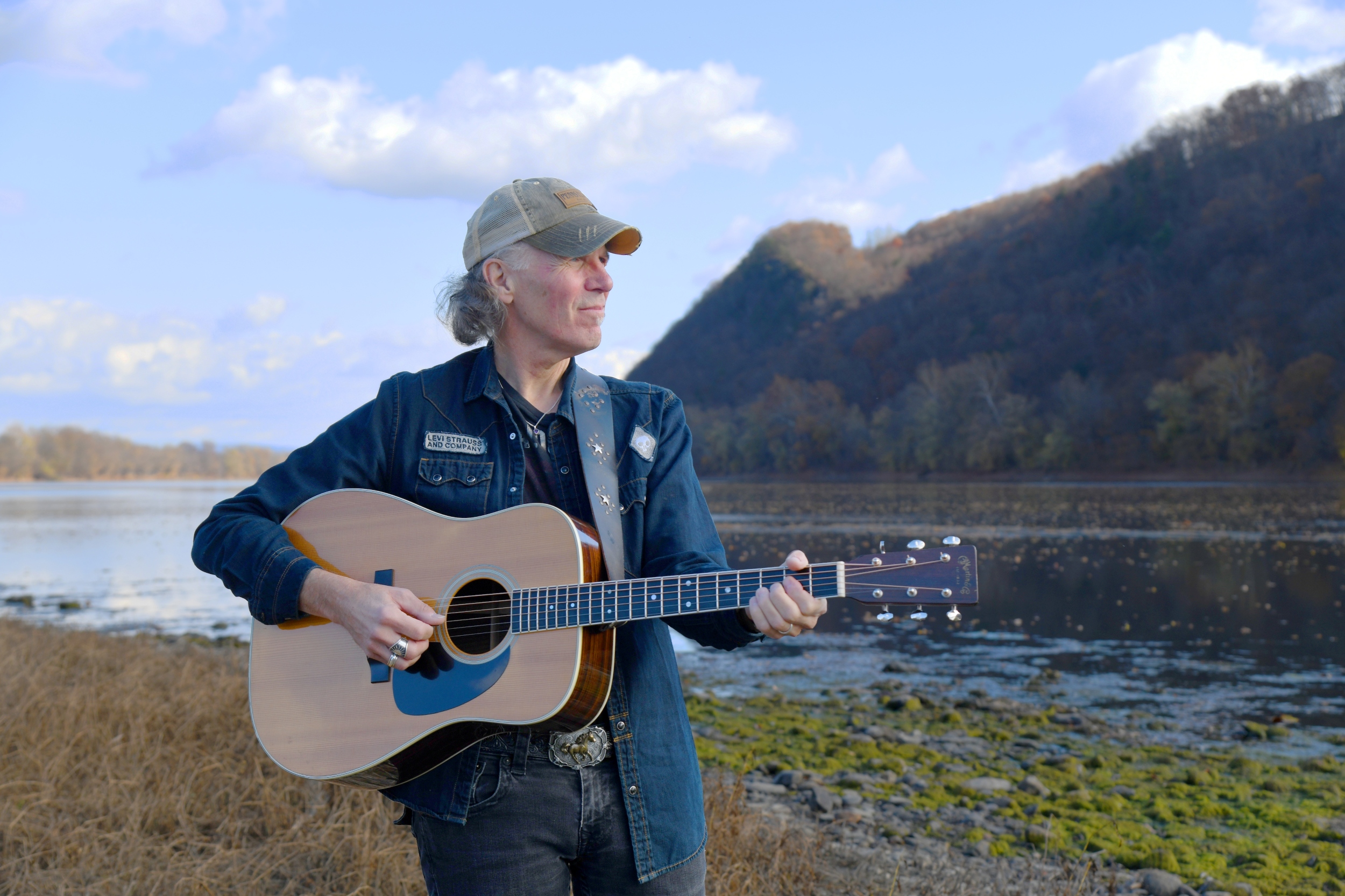 Brooks playing his guitar by a river with trees and hill behind