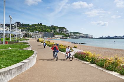 Two men cycling towards the camera on Dover Promenade with colourful flowers in beds, Dover Harbour, Dover Castle and the White Cliffs in the background