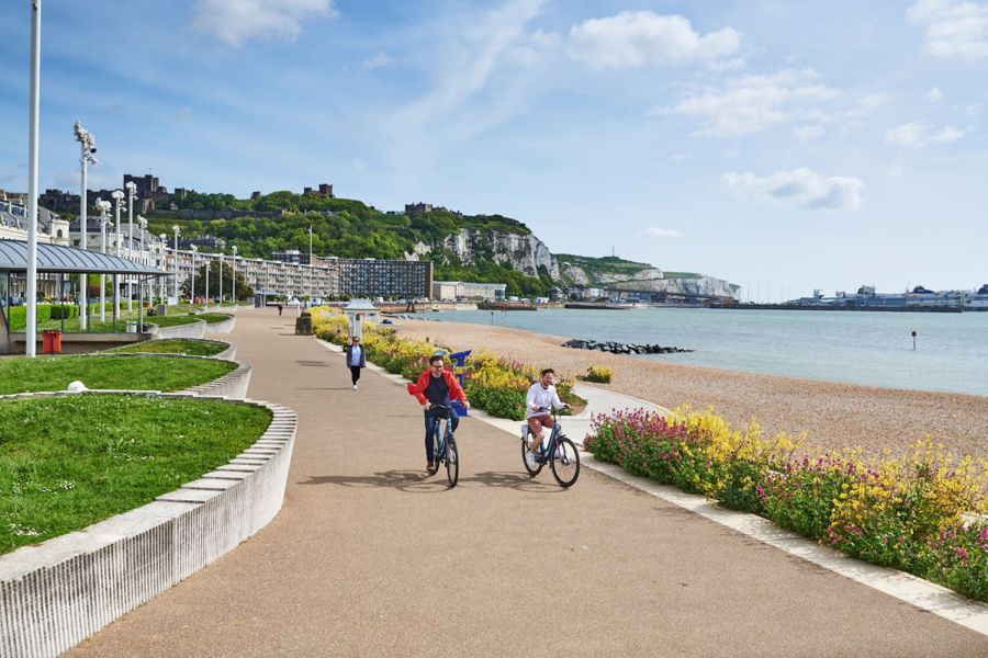 Two men cycling towards the camera along Dover Seafront with the smart architecture of the promenade clearly visible, wildflowers and sea to the right and Dover Castle on the cliff in the distance.