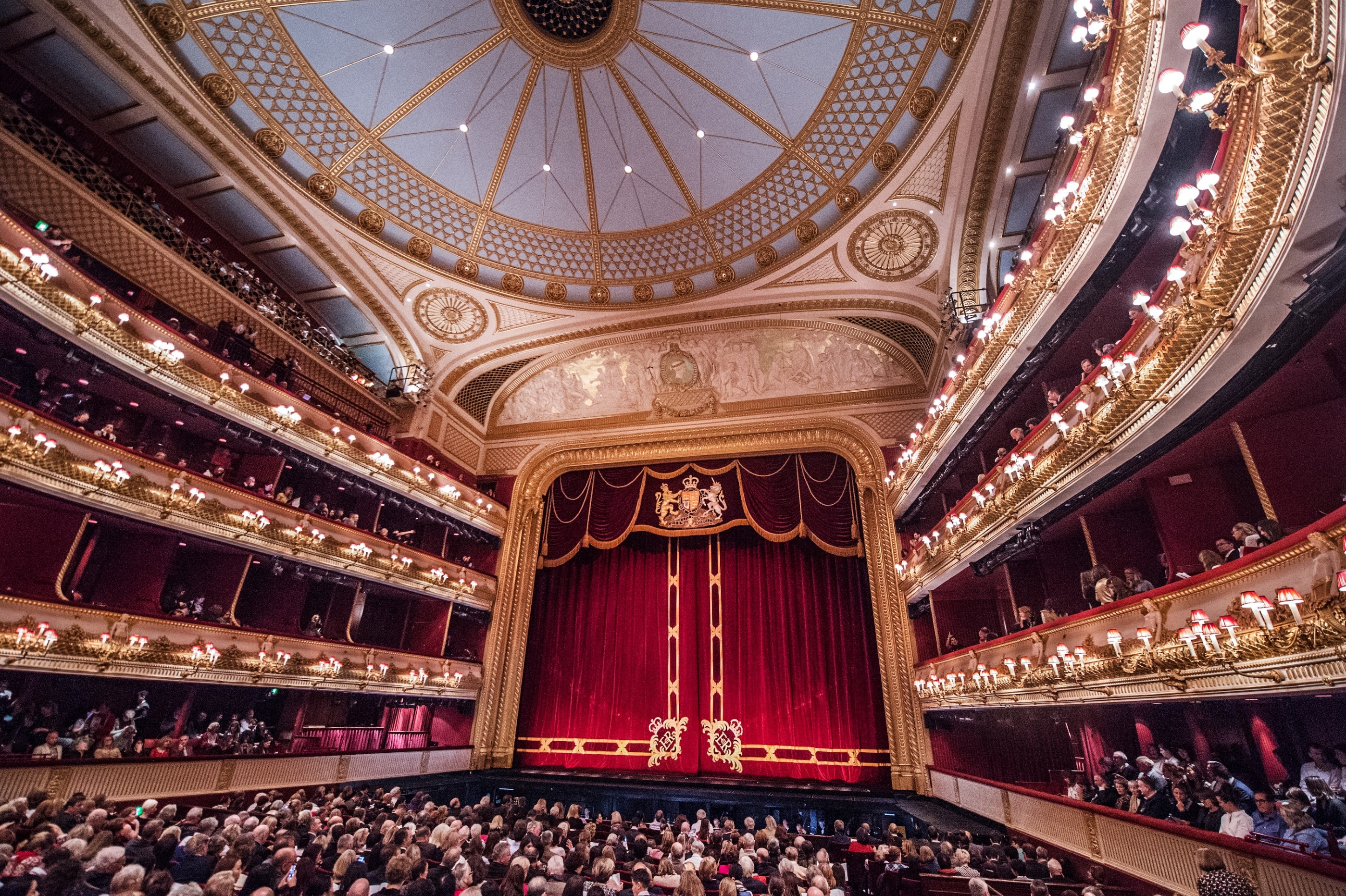An audience packed large theatre with closed red velvet curtains featuring the royal crest