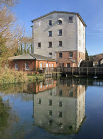 A five-storey timber-clad water mill and outbuildings reflected in a still millpond with a blue sky