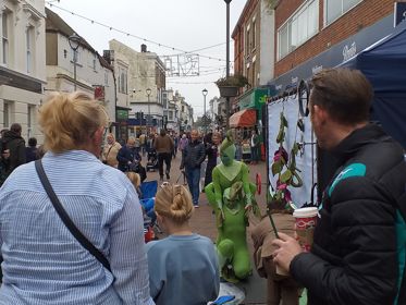 A busy high street with a street performer in a green outfit.