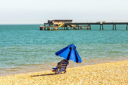 A single blue and white striped deckchair on a pebbly beach with a blue umbrella and Deal Pier in the background.