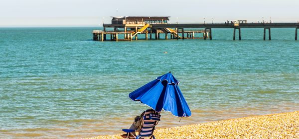 A single blue and white striped deckchair on a pebbly beach with a blue umbrella and Deal Pier in the background.