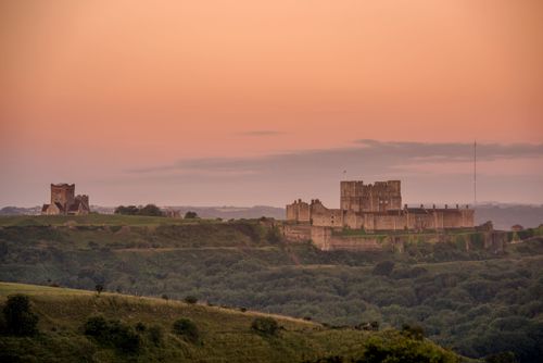 Dover Castle from a distance in the early morning light with a pink sky and greenery in the foreground.