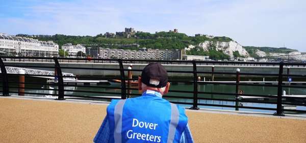 A man wearing a blue high-vis tabard with the words Dover Greeters on the back sitting on a bench in the foreground, with Dover seafront and castle in the distance
